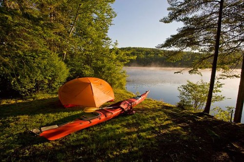 A bright orange tent and a red kayak by a serene lake surrounded by lush green trees at sunrise.