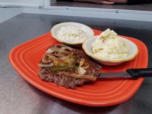 A vibrant orange plate with a grilled burger topped with onions and peppers, accompanied by two bowls of coleslaw and potato salad.