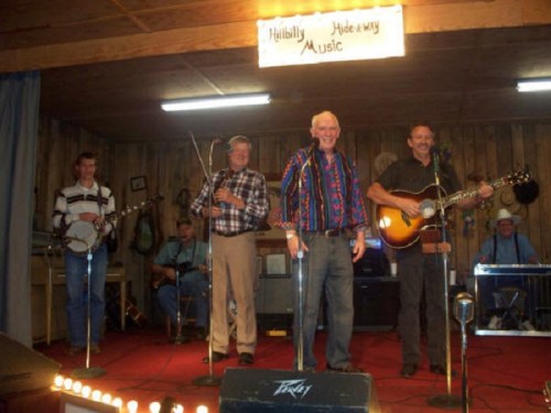 Four musicians perform on stage at a venue, with instruments like a banjo and guitar, under a sign reading "Hillbilly Hideaway Music."