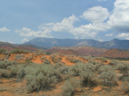 A scenic view of mountains in the background with sandy hills and sparse vegetation in the foreground under a cloudy sky.
