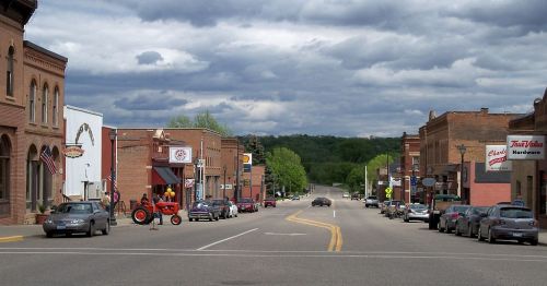 A small town street lined with shops, parked cars, and a tractor, under a cloudy sky.