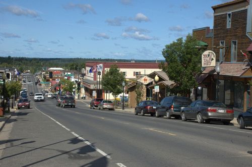 A view of a small town street lined with shops and parked cars under a blue sky with scattered clouds.