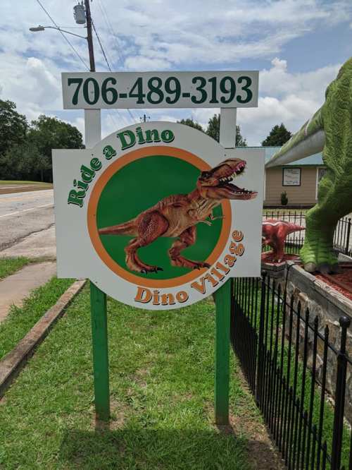 Sign for "Dino Village" featuring a dinosaur graphic and contact number, set against a blue sky and greenery.