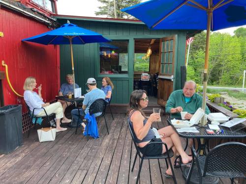 A group of people dining outdoors on a deck, enjoying food and drinks under blue umbrellas.