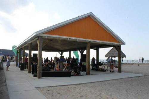 A beachside pavilion with people gathered, featuring a sandy area and a clear sky in the background.
