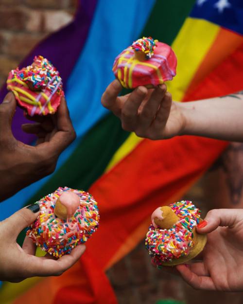 Four hands hold colorful donuts with sprinkles against a vibrant rainbow flag backdrop.