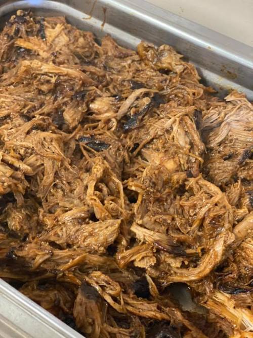 A close-up of shredded, cooked meat in a metal tray, glistening with a rich, dark sauce.