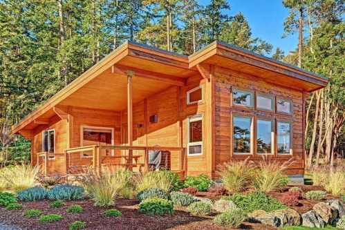 A modern wooden cabin surrounded by lush greenery and colorful landscaping under a clear blue sky.