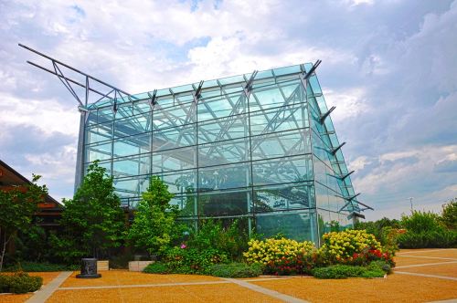 A modern glass building surrounded by greenery and colorful flowers under a cloudy sky.