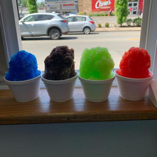 Four colorful snow cones in white cups sit on a windowsill, with blue, brown, green, and red flavors visible.