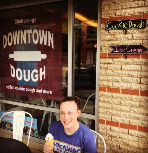 A person smiles while enjoying ice cream outside a shop with signs for cookie dough and ice cream in downtown Chattanooga.