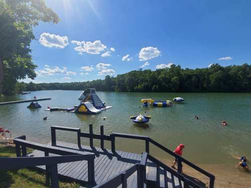 A sunny lakeside scene with inflatable water slides and people swimming, surrounded by trees and blue skies.