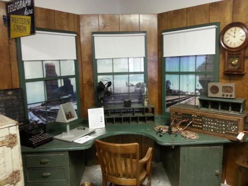 A vintage office setup with a wooden desk, typewriter, fan, and old telecommunication equipment against a wooden wall.