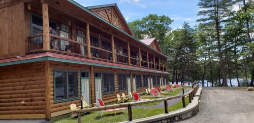 A wooden lodge with balconies, red chairs outside, and trees in the background near a lake on a sunny day.
