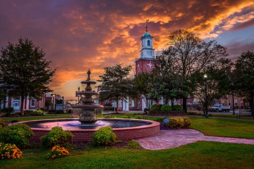 A scenic sunset over a town square featuring a fountain, trees, and a historic building with a clock tower.