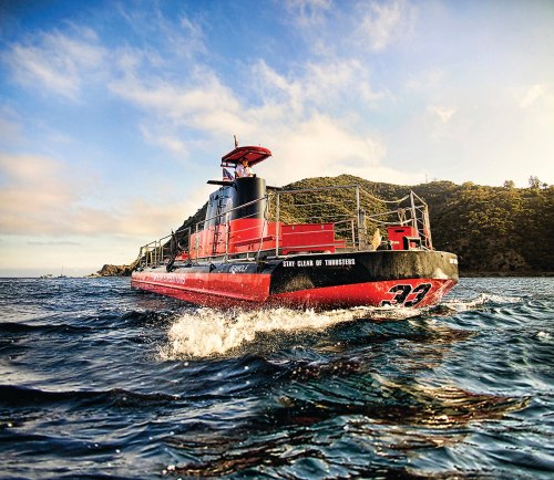 A red and black boat navigates through water, with a hilly coastline in the background under a blue sky.