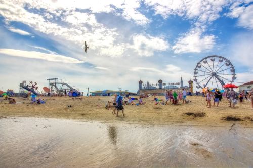 A lively beach scene with people, colorful umbrellas, a ferris wheel, and amusement rides under a blue sky.