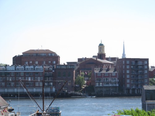 View of a riverfront cityscape featuring buildings, a clock tower, and a church steeple against a clear blue sky.