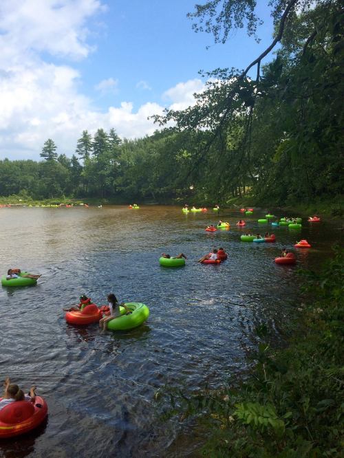 Colorful inner tubes float on a calm river, with people relaxing and enjoying a sunny day surrounded by greenery.