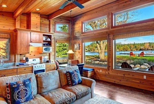 Cozy wooden cabin interior with a kitchen, sofa, and large windows showcasing a scenic lake view.