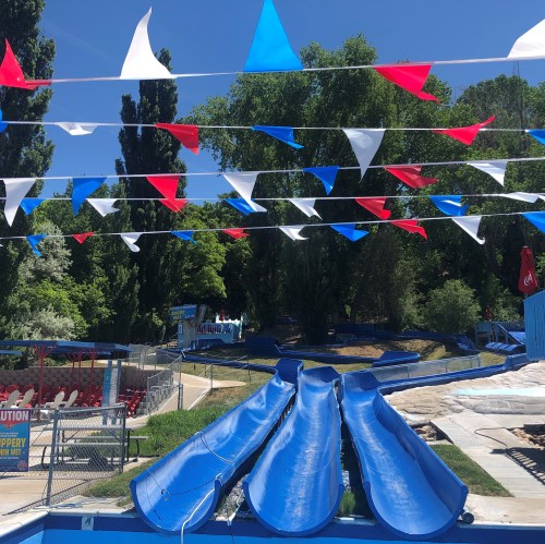 Colorful flags hang above blue water slides at a sunny water park, surrounded by green trees and empty seating.