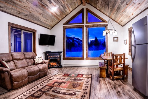 Cozy cabin interior with a brown sofa, wooden dining table, and large windows showcasing a snowy mountain view.