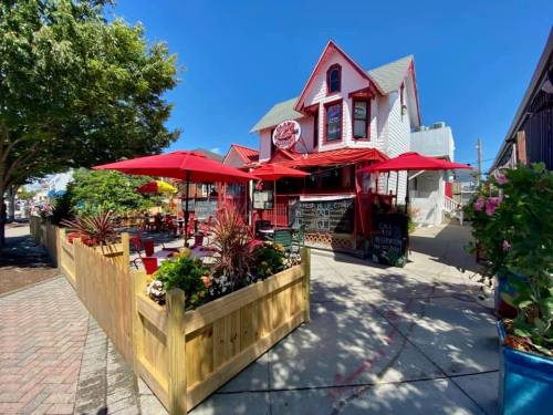 A charming restaurant with red umbrellas, surrounded by greenery and a wooden fence, under a clear blue sky.