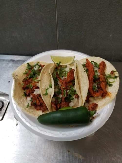 Three tacos filled with seasoned meat, onions, and cilantro, served with a lime wedge and a jalapeño on the side.