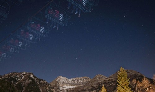 A night sky filled with stars above a mountain landscape, featuring a ski lift in the foreground.