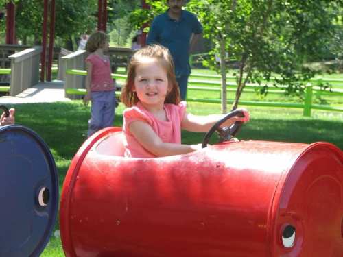 A young girl in a pink shirt smiles while sitting in a red toy car at a park, surrounded by greenery.
