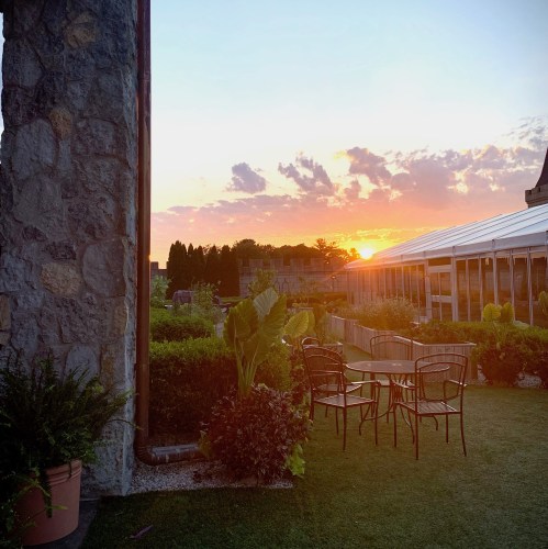 A sunset view from a rooftop garden, featuring chairs, greenery, and a stone wall against a colorful sky.