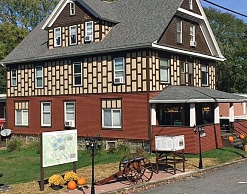 A large, two-story house with a unique brown and tan exterior, surrounded by greenery and pumpkins on the ground.