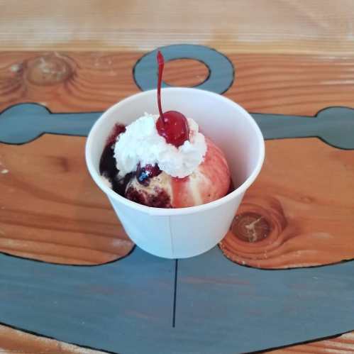 A cup of ice cream topped with whipped cream and a cherry, placed on a wooden table with a blue anchor design.