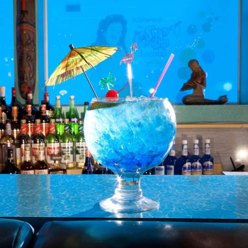A vibrant blue cocktail in a large glass, garnished with a cherry, umbrella, and flamingo, set against a bar backdrop.