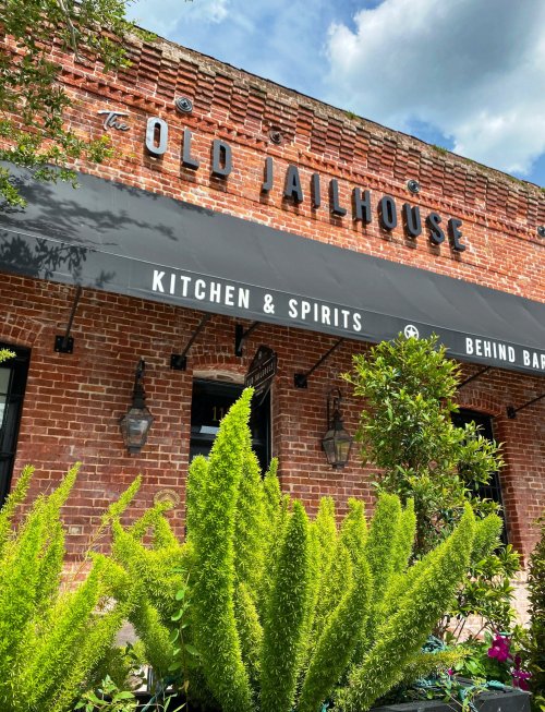 Exterior of The Old Jailhouse restaurant, featuring a brick facade, awning, and lush green plants in front.