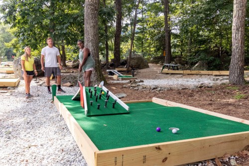 Three people play mini-golf on a green course surrounded by trees, with one preparing to take a shot.
