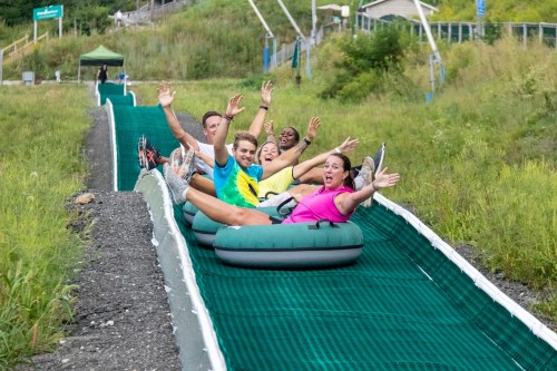 A group of six people joyfully sliding down a green tubing slope, hands raised in excitement.