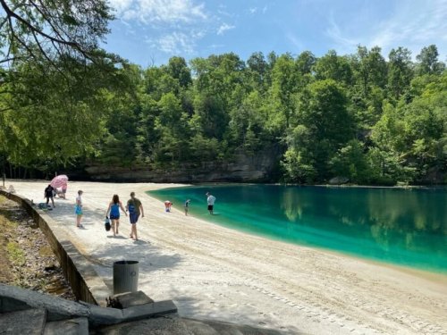 A sandy beach by a clear blue lake, surrounded by lush green trees, with people walking and enjoying the scenery.