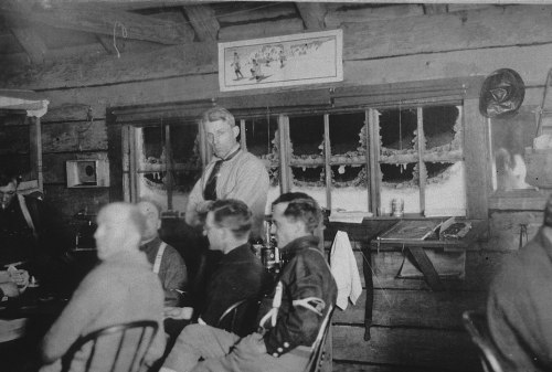 A black-and-white photo of men seated at a table in a rustic interior, with one man standing by a window.