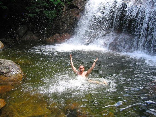 A person joyfully splashes in a natural pool beneath a waterfall, surrounded by rocks and greenery.