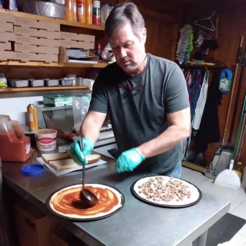 A man in a gray shirt and gloves spreads sauce on a pizza crust in a kitchen, with toppings on a separate plate.