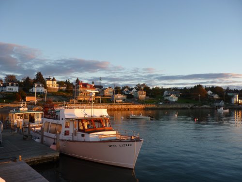 A boat named "Miss Lizzie" docked at a serene harbor with colorful houses and a sunset sky in the background.