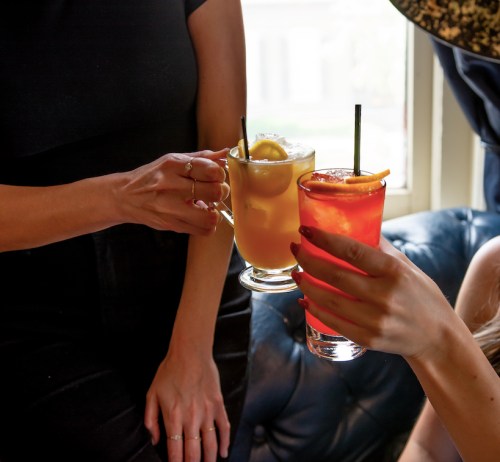 Two hands clink glasses of colorful cocktails, one with a lemon slice and the other with a slice of orange.