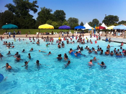 A crowded outdoor pool filled with people enjoying the water on a sunny day, with colorful umbrellas in the background.