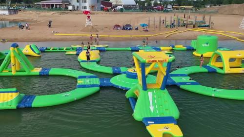 A colorful inflatable water park with slides and obstacles, set on a lake with sandy beach and people enjoying the activities.