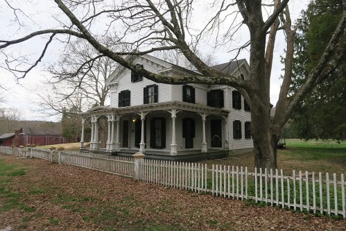 A white Victorian-style house with black shutters, surrounded by a picket fence and bare trees in a rural setting.