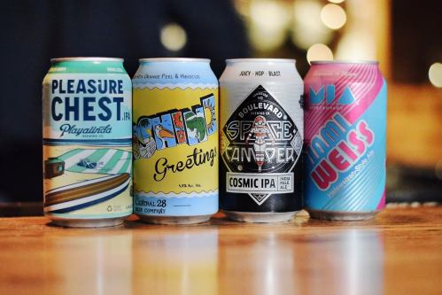 Four colorful beer cans lined up on a wooden surface, featuring various designs and labels for different IPAs.
