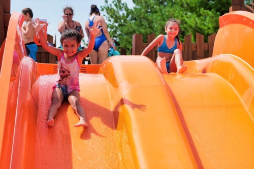Two children joyfully slide down bright orange water slides on a sunny day, with other people in the background.
