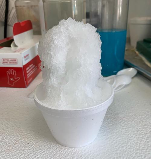 A white cup filled with fluffy, crushed ice, resembling a snow cone, on a textured surface.