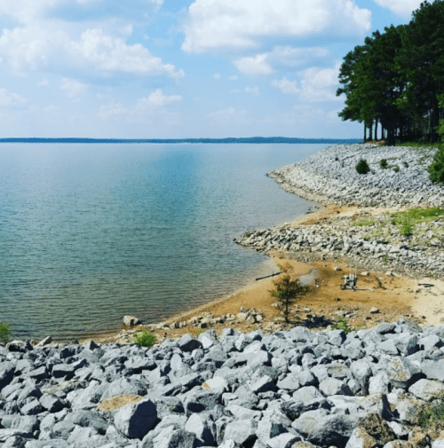 A serene lakeside view with rocky shorelines, clear water, and a blue sky dotted with clouds.